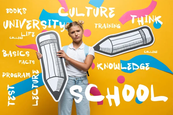 Sad schoolgirl holding paper pencil and looking at camera near lettering on yellow background — Stock Photo