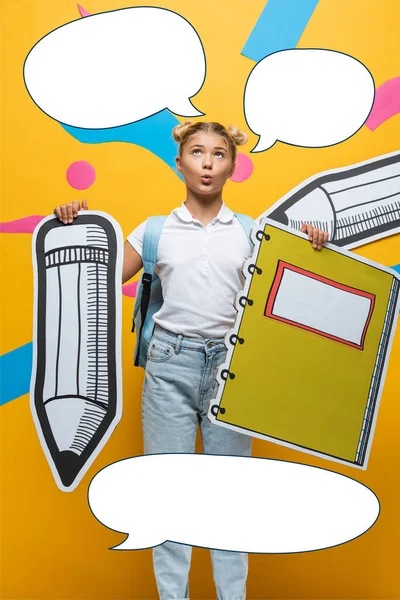 Pensive школярка тримає паперовий олівець і блокнот біля ілюстрації бульбашок мовлення на жовтому — стокове фото