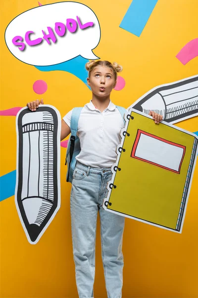 Colegiala pensativa sosteniendo lápiz de papel y cuaderno cerca de la ilustración de la burbuja del habla con letras de la escuela en amarillo - foto de stock