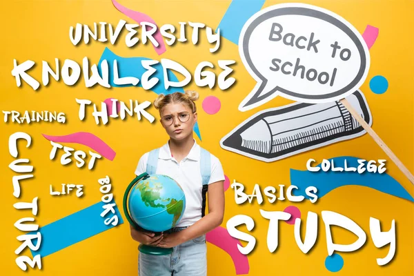 Thoughtful schoolkid with globe standing beside paper art with back to school lettering and illustration on yellow — Stock Photo