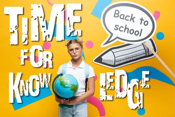 Écolier réfléchi avec globe debout à côté de l'art du papier avec retour à l'école lettrage près de l'illustration sur fond jaune — Photo de stock
