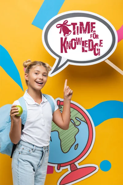 Schoolkid with backpack and apple pointing at paper speech bubble with time for knowledge lettering on yellow — Stock Photo