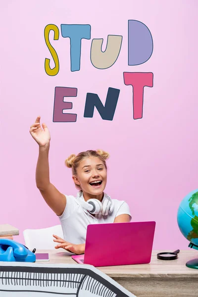 Schoolgirl sitting with raised hand near laptop, telephone, student lettering and paper art on pink — Stock Photo