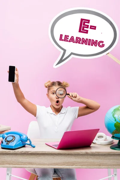 Scolaretta con lente d'ingrandimento e smartphone con schermo bianco vicino alla bolla vocale con lettering e-learning, globo e laptop su rosa — Foto stock