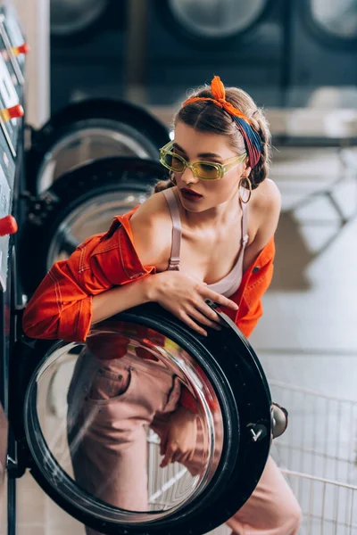Mulher elegante em óculos de sol olhando para a máquina de lavar roupa na lavanderia — Fotografia de Stock