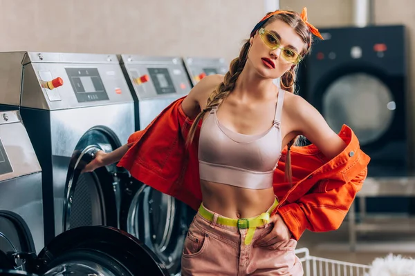 Stylische Frau mit Sonnenbrille blickt in die Kamera und steht neben Waschmaschinen im Waschsalon — Stockfoto