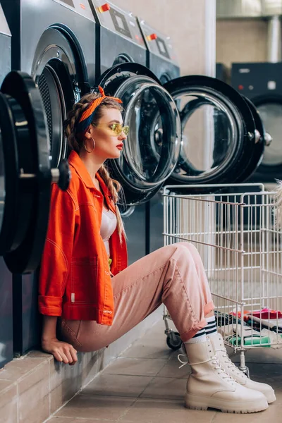 Stylish woman in sunglasses sitting near metallic cart and washing machines in laundromat — Stock Photo