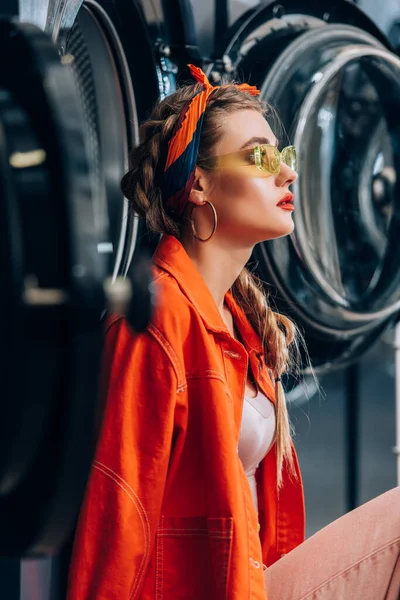 Mujer con estilo en gafas de sol sentado cerca de las lavadoras en la lavandería con el primer plano borroso - foto de stock