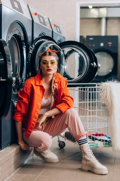 Stylish woman in sunglasses sitting near cart with dirty clothing and washing machines in laundromat — Stock Photo