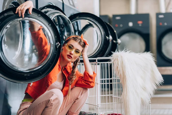 Trendy woman in sunglasses sitting near metallic cart and washing machines in laundromat — Stock Photo