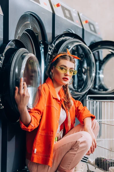 Mujer de moda en gafas de sol sentado cerca de carro metálico y lavadoras en lavandería - foto de stock