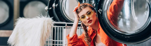 Trendy woman in sunglasses sitting near metallic cart with faux fur and washing machines in laundromat, banner — Stock Photo