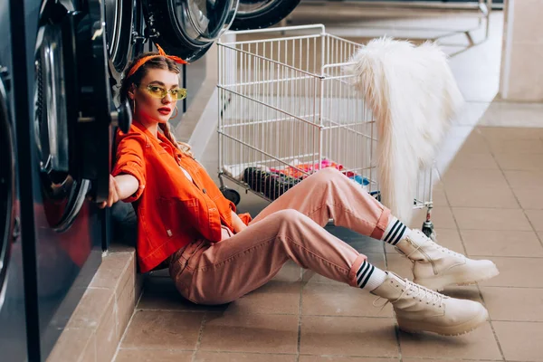 Mujer de moda en gafas de sol sentada en el suelo cerca de carro metálico con ropa sucia y lavadoras en lavandería - foto de stock