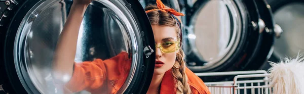 Молода жінка в сонцезахисних окулярах дивиться на камеру через двері пральної машини, банер — стокове фото