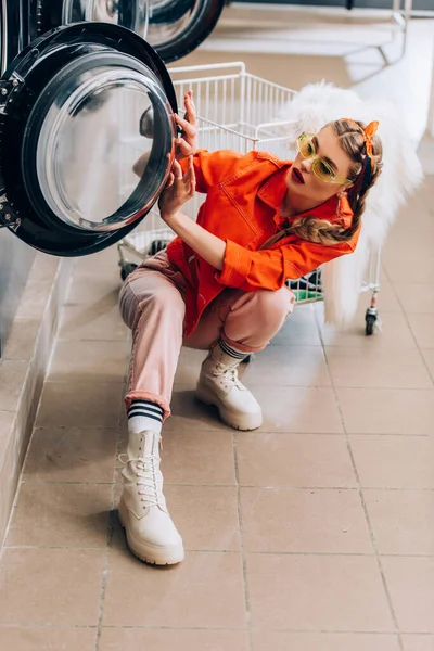 Mujer con estilo en gafas de sol mirando a la lavadora moderna en lavandería - foto de stock