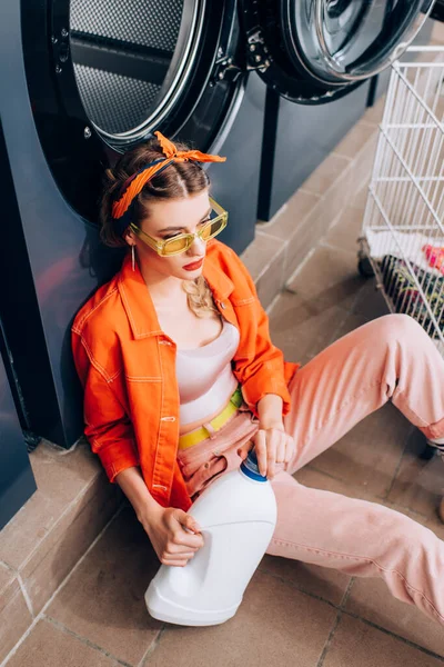 High angle view of stylish woman in sunglasses sitting on floor and holding bottle with detergent near washing machine in laundromat — Stock Photo