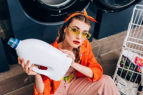 High angle view of young woman sitting on floor and holding bottle with detergent in laundromat — Stock Photo