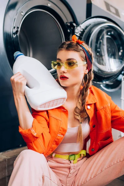 Jeune femme en lunettes de soleil tenant bouteille avec détergent dans la laverie automatique — Photo de stock