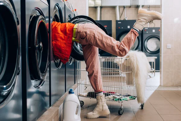 Mujer joven revisando el interior de la lavadora cerca de la botella de detergente y el carro con ropa sucia - foto de stock