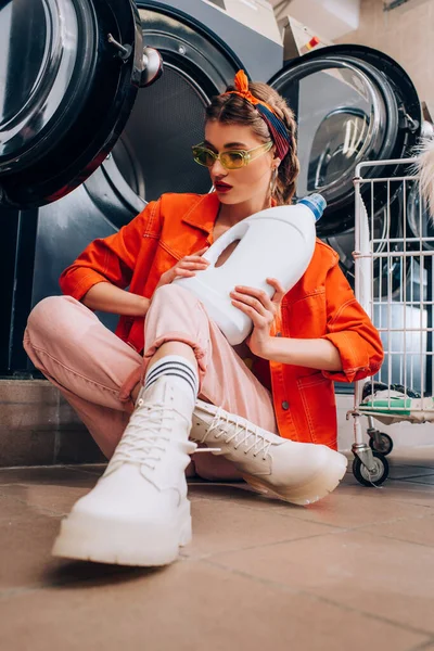 Mujer joven en gafas de sol sosteniendo botella con detergente y sentada en el suelo en la lavandería - foto de stock