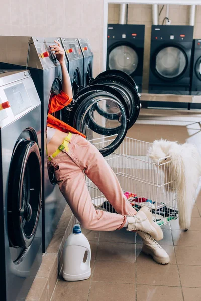 Piernas de mujer curiosa que sobresalen de la lavadora cerca de la botella de detergente y el carro con ropa sucia - foto de stock