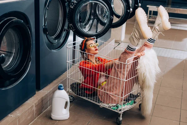 Stylish woman sitting in cart near washing machines and bottle with detergent in laundromat — Stock Photo