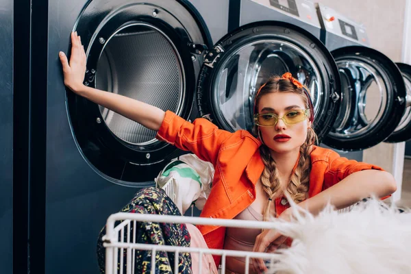 Stylish woman sitting in cart with clothing near washing machines in laundromat — Stock Photo