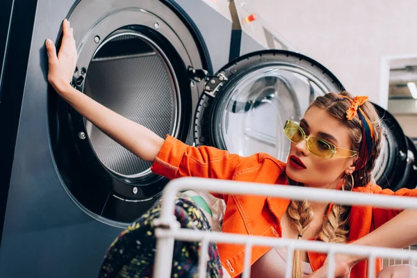 Stilvolle Frau sitzt in Einkaufswagen neben Waschmaschinen im Waschsalon mit verschwommenem Vordergrund — Stockfoto