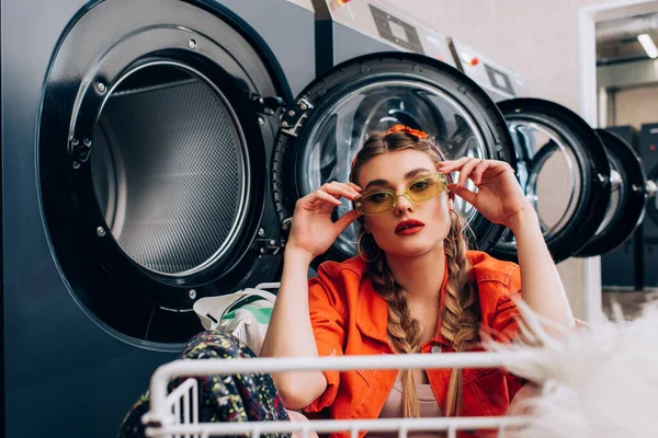 Stylish woman touching sunglasses and sitting in cart near washing machines in laundromat — Stock Photo
