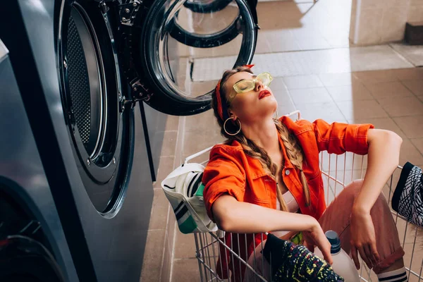 Stylish woman sitting in cart with clothing and detergent near washing machines in laundromat — Stock Photo