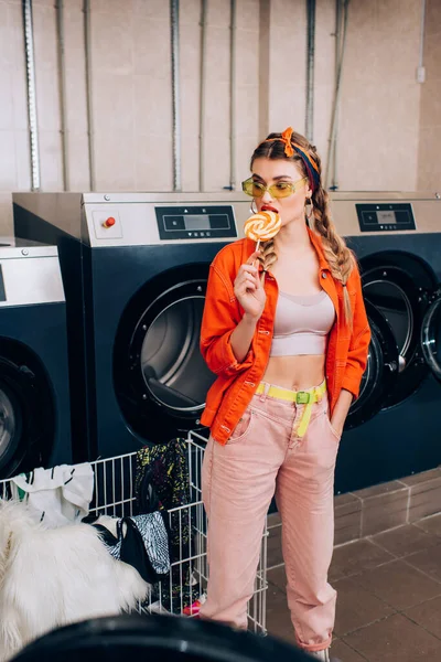 Junge stylische Frau leckt Lutscher in der Nähe von Einkaufswagen mit Kleidung und Waschmaschinen im Waschsalon — Stockfoto