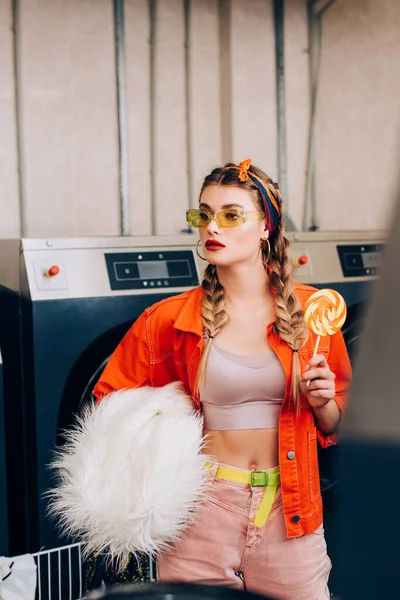 Young stylish woman holding lollipop and faux fur coat near washing machines in laundromat — Stock Photo