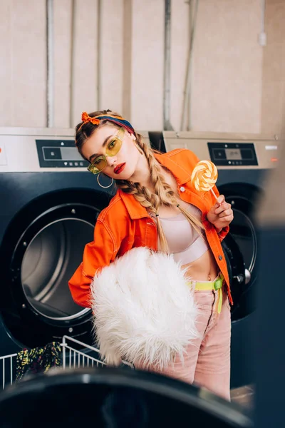Mulher elegante em óculos de sol segurando pirulito e casaco de peles falso perto de máquinas de lavar roupa na lavanderia — Fotografia de Stock