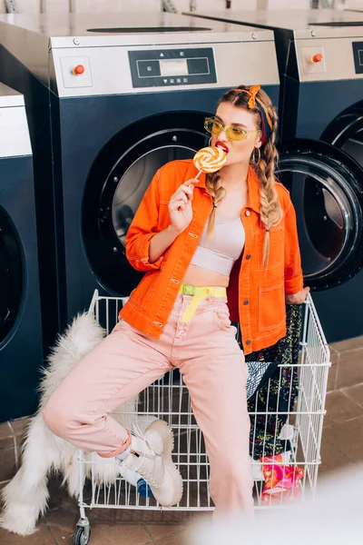 Stilvolle Frau mit Sonnenbrille beißt Lutscher neben Einkaufswagen mit Kleidung und Waschmaschinen im Waschsalon — Stockfoto