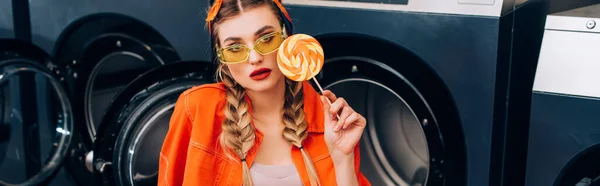 Stylish woman in sunglasses holding lollipop near washing machines in laundromat, banner — Stock Photo