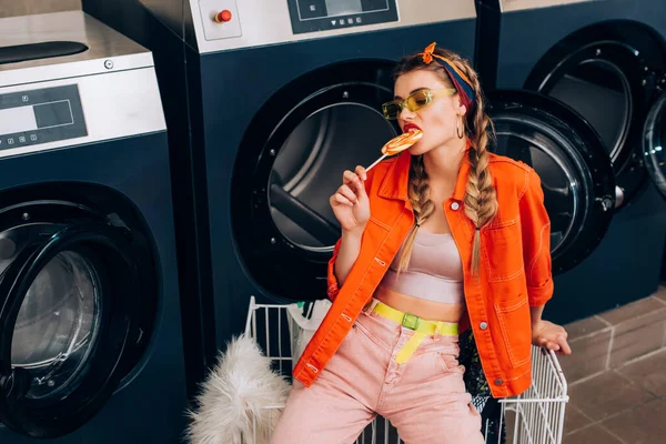 Femme à la mode dans les lunettes de soleil mordant sucette près du chariot et machines à laver dans la laverie automatique — Photo de stock