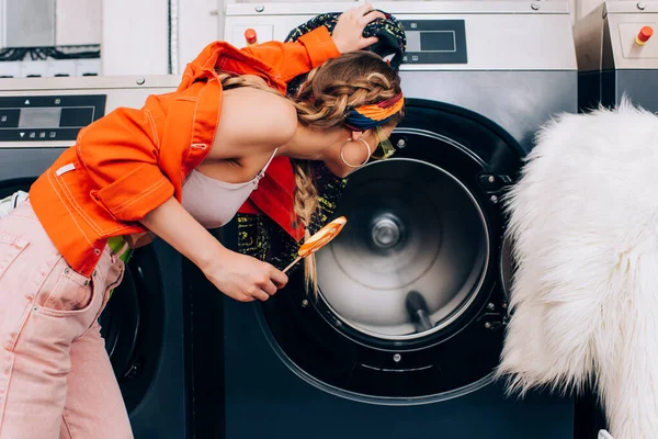 Trendy young woman in sunglasses holding lollipop and checking washing machine in laundromat — Stock Photo