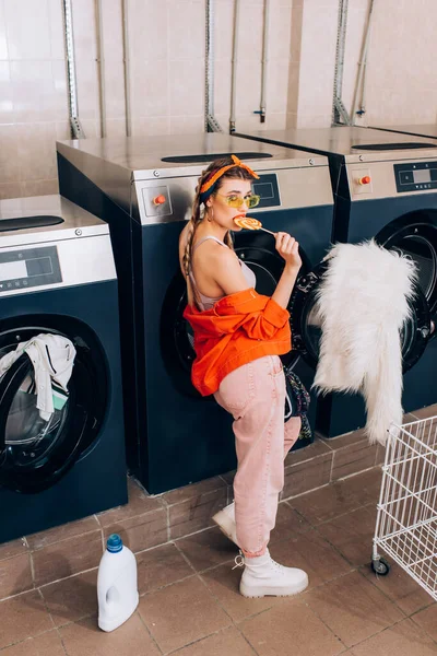 Mujer joven de moda en gafas de sol con piruleta cerca de lavadoras en lavandería - foto de stock