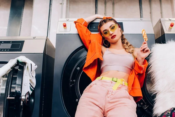 Fashionable woman in sunglasses holding lollipop in modern laundromat — Stock Photo