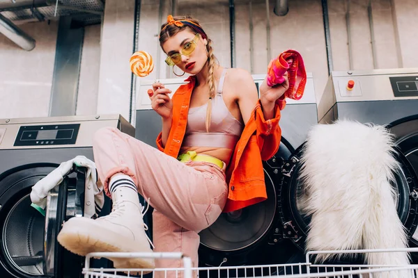 Fashionable woman in sunglasses holding sweet lollipop in modern laundromat with blurred foreground — Stock Photo