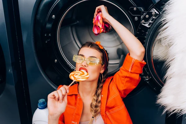 Femme à la mode mordre sucette douce et tenant des vêtements dans la laverie automatique — Photo de stock