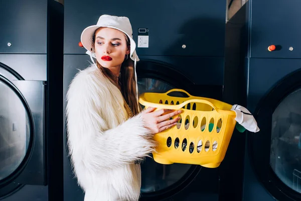 Stylish woman in faux fur jacket and hat holding basket with clothing near washing machines in laundromat — Stock Photo