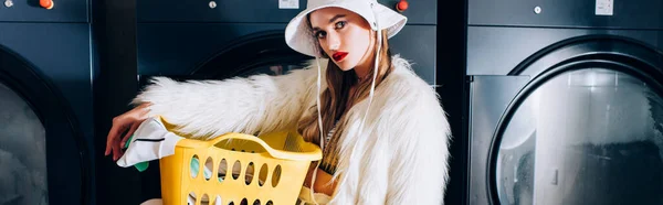 Stylish woman in faux fur jacket and hat holding basket with laundry near washing machines in laundromat, banner — Stock Photo