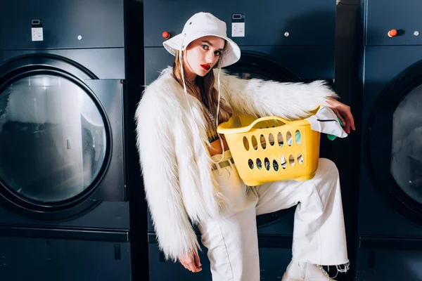 Stylish woman in faux fur jacket and hat holding basket with laundry near washing machines in laundromat — Stock Photo