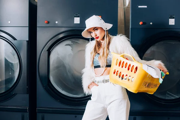 Stylish woman in faux fur jacket and hat holding basket with laundry and standing with hand in pocket near washing machines in laundromat — Stock Photo