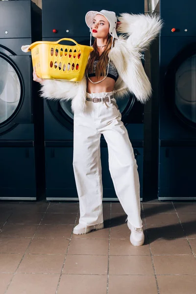 Jovem mulher em casaco de pele falso e chapéu segurando cesta com lavanderia perto de máquinas de lavar roupa na lavanderia — Fotografia de Stock