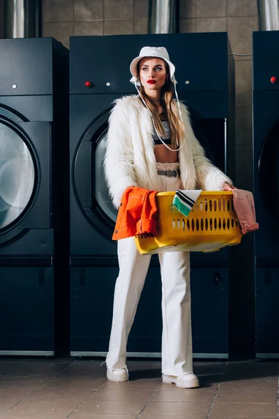 Élégant jeune femme en fausse veste de fourrure et chapeau tenant panier avec buanderie près des machines à laver dans la laverie automatique — Photo de stock