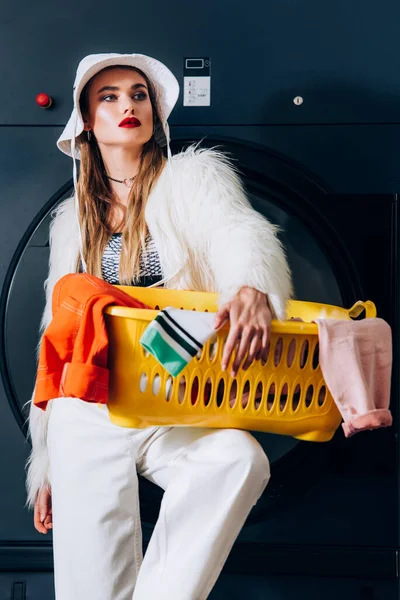 Jovem na moda em casaco de pele falso e chapéu segurando cesta com lavanderia perto da máquina de lavar roupa na lavanderia — Fotografia de Stock