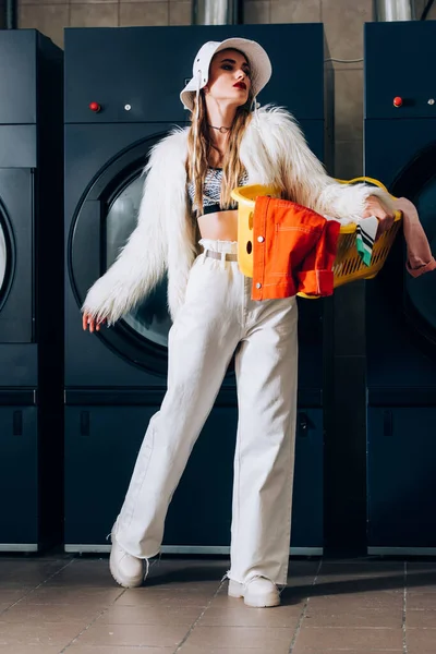 Femme élégante en fausse veste de fourrure et chapeau tenant panier avec buanderie près des machines à laver dans la laverie automatique — Photo de stock