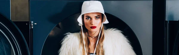 Elegante giovane donna in pelliccia sintetica giacca e cappello vicino alla lavatrice in lavanderia, banner — Foto stock